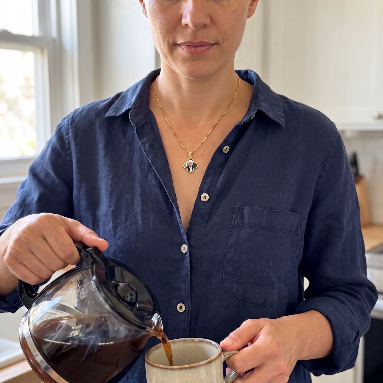Woman in a navy linen shirt pouring coffee in her kitchen, wearing the gold secret clover photo necklace | SKU: TPH-CLVN-GOLD