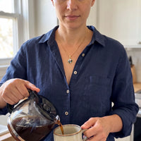 Thumbnail for Woman in a navy linen shirt pouring coffee in her kitchen, wearing the gold secret clover photo necklace | SKU: TPH-CLVN-GOLD